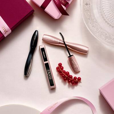 Flat-lay aranžman Lancome proizvoda za šminku, s maskarom i sjajilom za usne, uz ružičaste poklon kutije, satenske vrpce i grančicu crvenih ribizli.