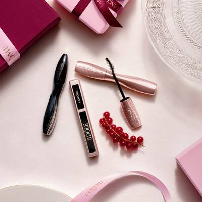 Prikaz Lancome make-up proizvoda iz zraka, uključujući maskaru i sjajilo za usne, uz ružičaste poklon-kutije, satenske vrpce i grančicu crvenog ribiza.