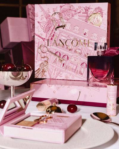 Svečani Lancome blagdanski postav s velikom ilustriranom ružičastom poklon-kutijom, parfemom i makeup proizvodima aranžiranim na stolu sa svježim trešnjama.