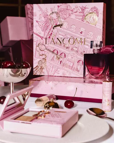 Svečani postav Lancome blagdanske linije na stolu, s velikom ilustriranom ružičastom poklon-kutijom, parfemom, makeup proizvodima i svježim trešnjama.