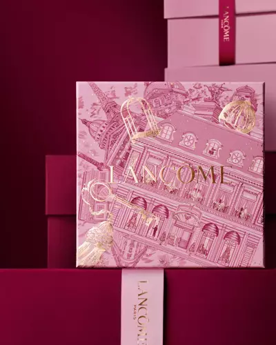 Slika ružičaste Lancome blagdanske poklon kutije koja stoji ispred hrpe ružičastih i tamnocrvenih poklon kutija, na tamnocrvenoj pozadini.