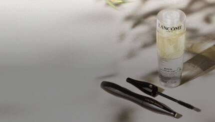 Na slici se nalazi proizvod za njegu kože "Lancome Clean & Care" u prozirnoj boci, uz crni tuš za oči "Lancome Hypnose", postavljen na reflektirajuću površinu, sa zelenim lišćem u pozadini.
