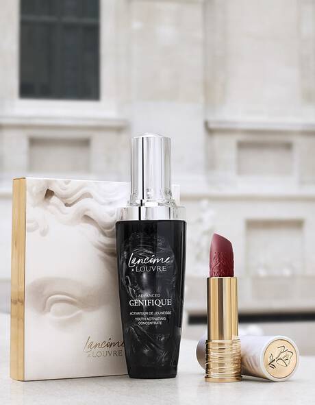 A bottle of Lancome advanced serum and a red lipstick sit next to a decorative box featuring a sculpture, with a blurred artistic background resembling the Louvre.