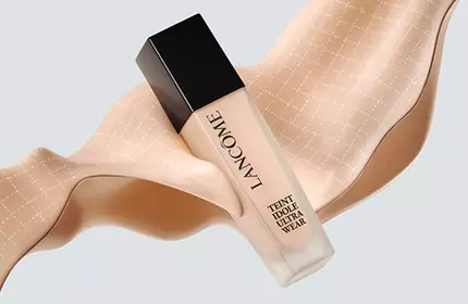 A bottle of foundation positioned on a flowing ribbon-like fabric in a soft beige tone, set against a light background.