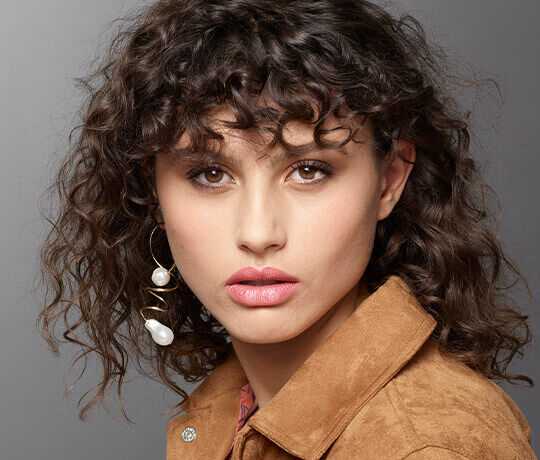A young woman with curly hair and bold makeup, wearing statement earrings and a suede jacket, gazes directly at the camera against a neutral background.