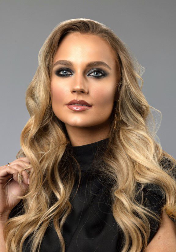 Žena s dugom, valovitom plavom kosom i sminkom u hladnim tonovima pozira s elegantnim izrazom lica.