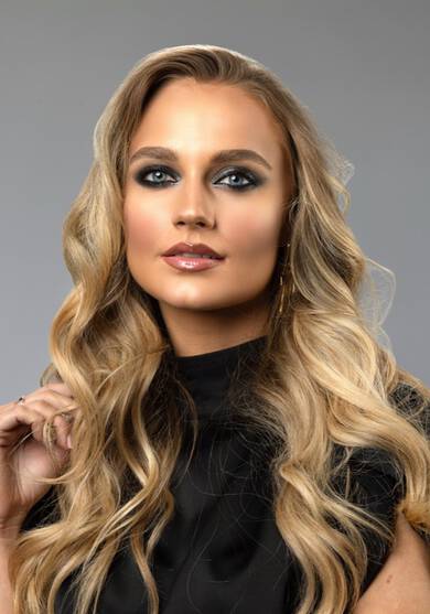 Žena s dugom, valovitom plavom kosom i sminkom u hladnim tonovima pozira s elegantnim izrazom lica.