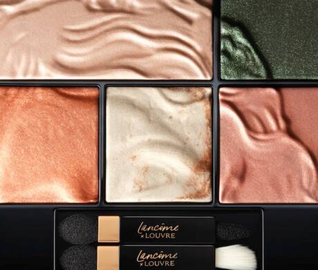 A close-up view of a makeup palette featuring various shimmering eyeshadow shades in warm and cool tones, arranged in distinct sections. Two double-ended applicators are included at the bottom.