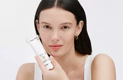 A woman with long, dark hair holds a skincare device while smiling at the camera against a light background.