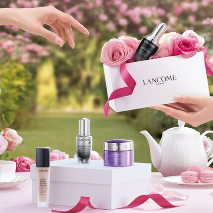 A beautifully arranged display featuring a gift box from Lancome filled with skincare products, surrounded by pink roses in a garden setting. Two hands are reaching for the gift, with a teapot and cups on a table nearby.