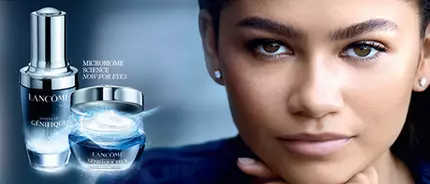 A close-up of a woman with dark hair and earrings, featuring skincare products from Lancome, including a serum and a cream, against a dark blue background.