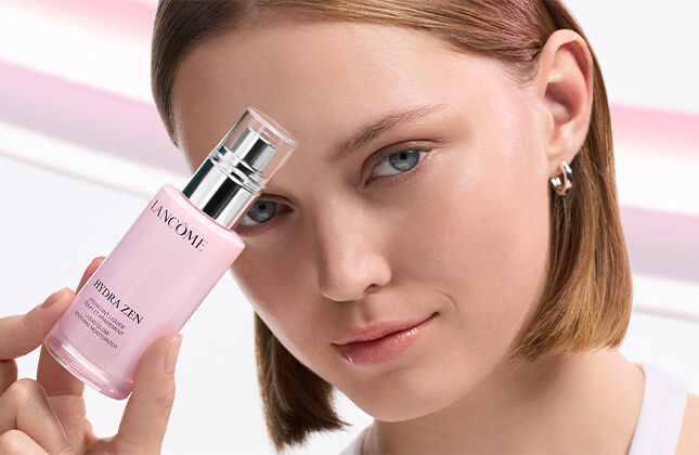 A young woman with short brown hair holds a pink skincare bottle close to her face, showcasing a fresh and radiant look against a soft, wavy background.