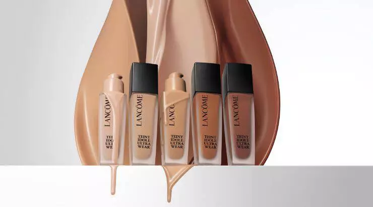 A collection of Lancome foundation bottles displayed in front of flowing liquid foundation swatches in various shades, showcasing the brand's Tient Idole Ultra Wear range.
