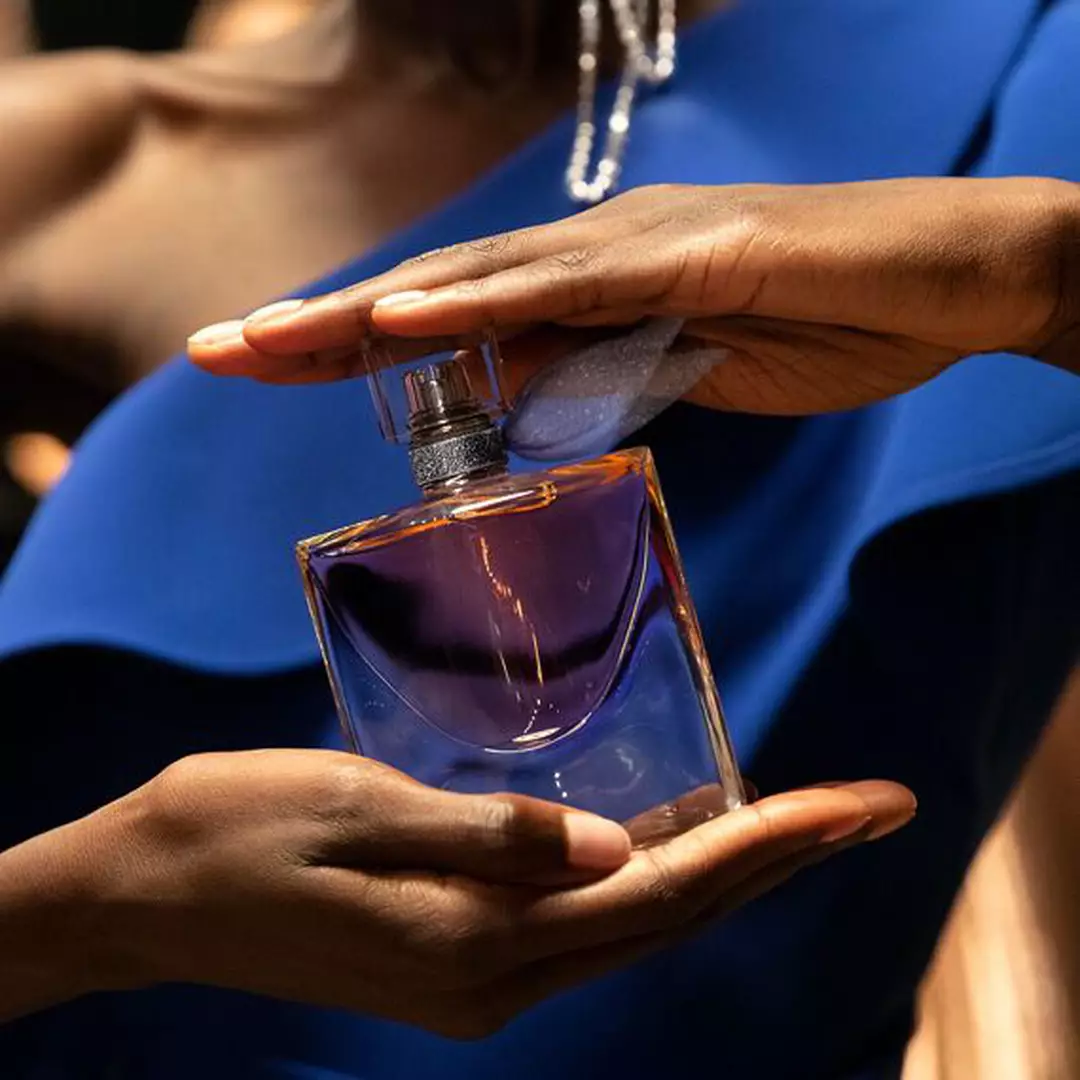 A close-up of a person holding a perfume bottle, with a soft focus on the elegant design and purple liquid inside. The individual wears a blue outfit and pearl accessories.