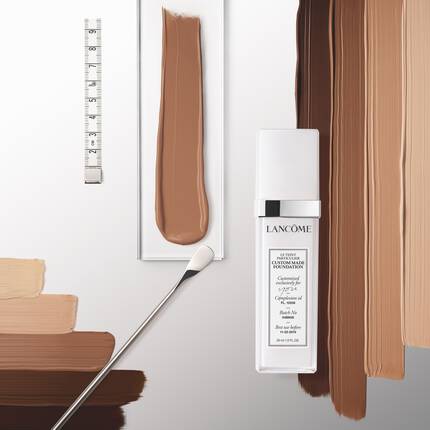 A bottle of Lancome skincare product displayed with various shades of foundation swatches arranged beside it on a sleek surface.