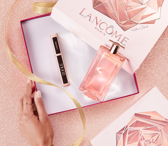 A hand reaches for a bottle of Lancome perfume and a lipstick inside an elegantly designed gift box, surrounded by gold ribbon on a shimmery pink surface.