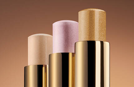Three makeup sticks in gold packaging arranged vertically on a gradient brown background. The sticks feature varying shades: light beige, soft pink, and a shimmer gold.