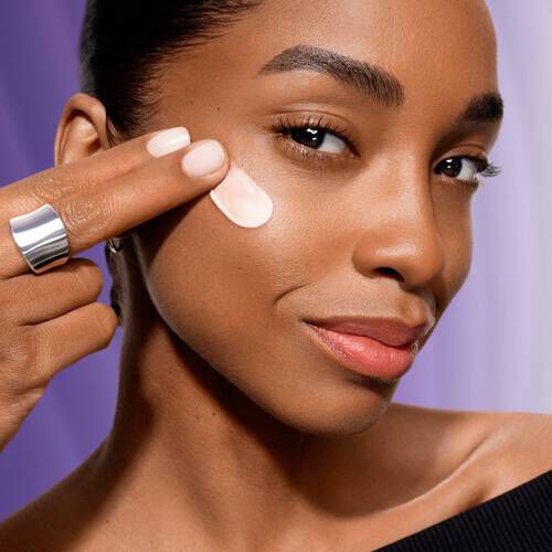 Žena s blistavom kožom nanosi kremu na obraz dok se smiješi pred ljubičastom pozadinom.