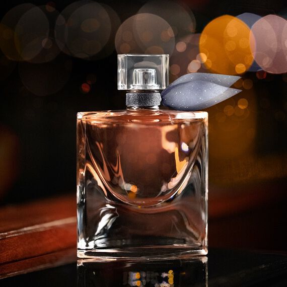 A stylish glass perfume bottle with a distinctive shape, featuring a silver cap and a decorative purple leaf on top, set against a blurred background with warm bokeh lights.