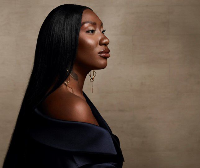 A profile portrait of a woman with long, straight black hair and dark skin, wearing an elegant off-shoulder top and statement earrings, set against a neutral background.