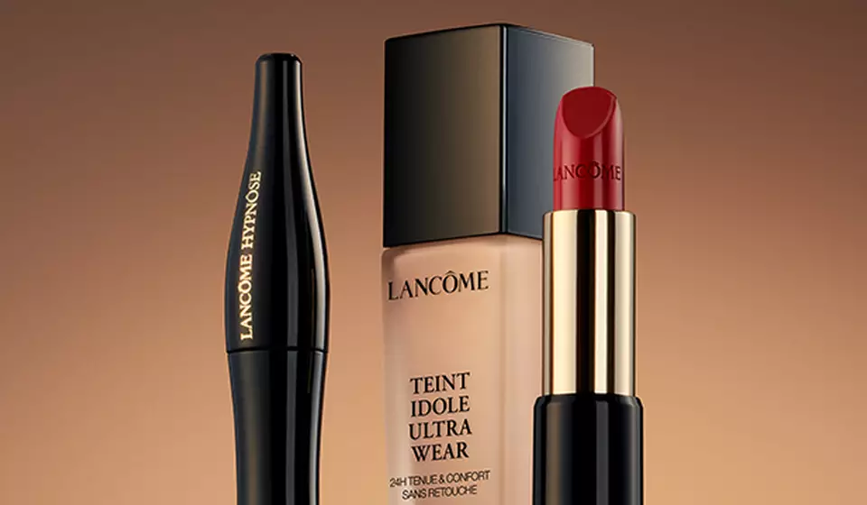 A close-up of three makeup products arranged elegantly: a black eyeliner pencil, a foundation bottle, and a red lipstick, all against a warm gradient background.