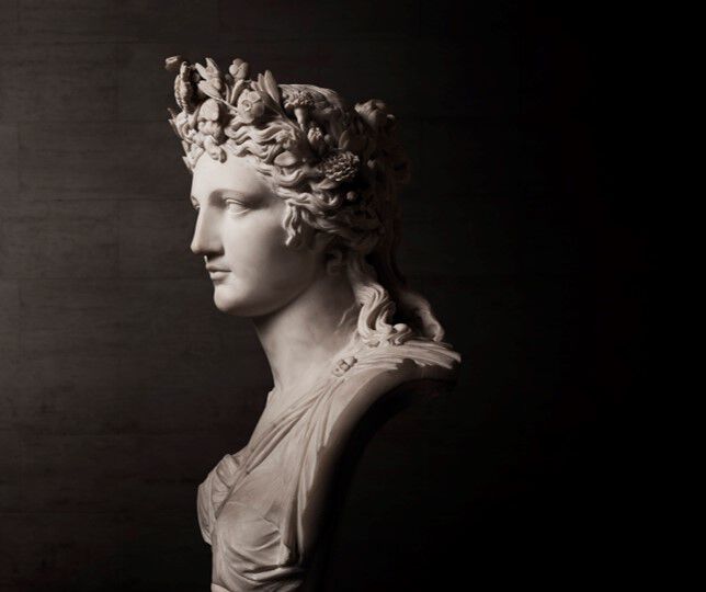 A side profile of a marble statue depicting a woman with a floral crown, elegantly draped attire, and detailed facial features, set against a dark background.