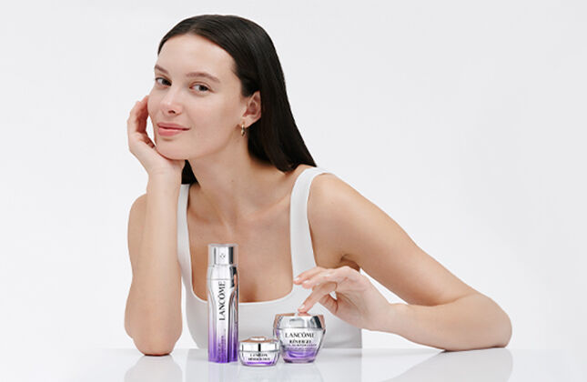 A woman with long dark hair is sitting at a table with skincare products in front of her, smiling gently at the camera. The background is minimalistic and soft in color.