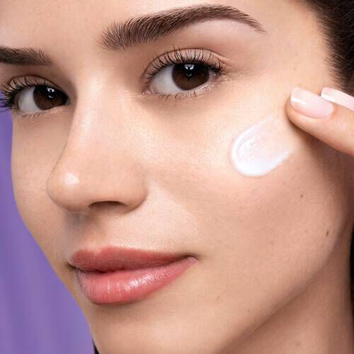 Mlada žena s prirodnim izgledom nanosi kremu na obraz ispod svjetlosne pozadine.