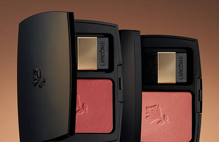 A close-up of two elegant black compact cases containing pink blushes, set against a warm, gradient background.