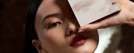 A close-up of a model's face, holding a beauty product close to her cheek. She has a bold lip color and subtle eye makeup, with a soft expression. The background is softly blurred.