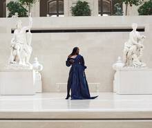 A model in a flowing navy gown stands elegantly on a marble platform surrounded by classical sculptures in a spacious, well-lit gallery.