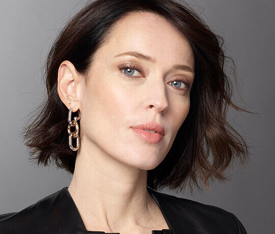 A close-up portrait of a woman with short dark hair, wearing a stylish black outfit and chic chain-link earrings. She has a natural makeup look, with soft, neutral tones on her lips and eyes.