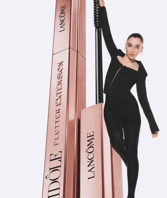 Žena u crnoj odjeći pozira uz dva pakiranja maskare Lancome Idole.