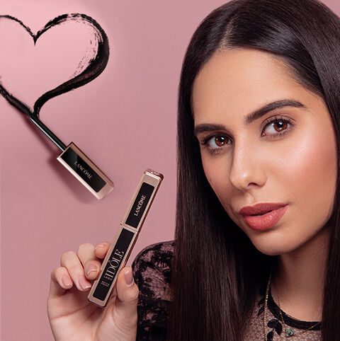 Žena s dugom tamnom kosom drži bočicu maskare ispred pozadine u ružičastim tonovima, s crnim srcom u lijevom kutu.